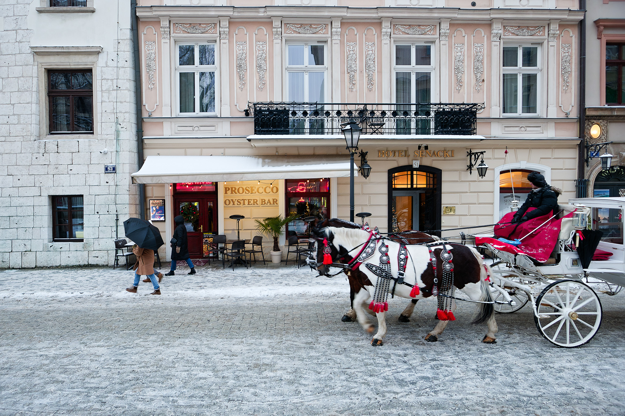 Zimowy Kraków – dorożka z końmi przed kamienicą i przechodnie z parasolami na krakowskim rynku.