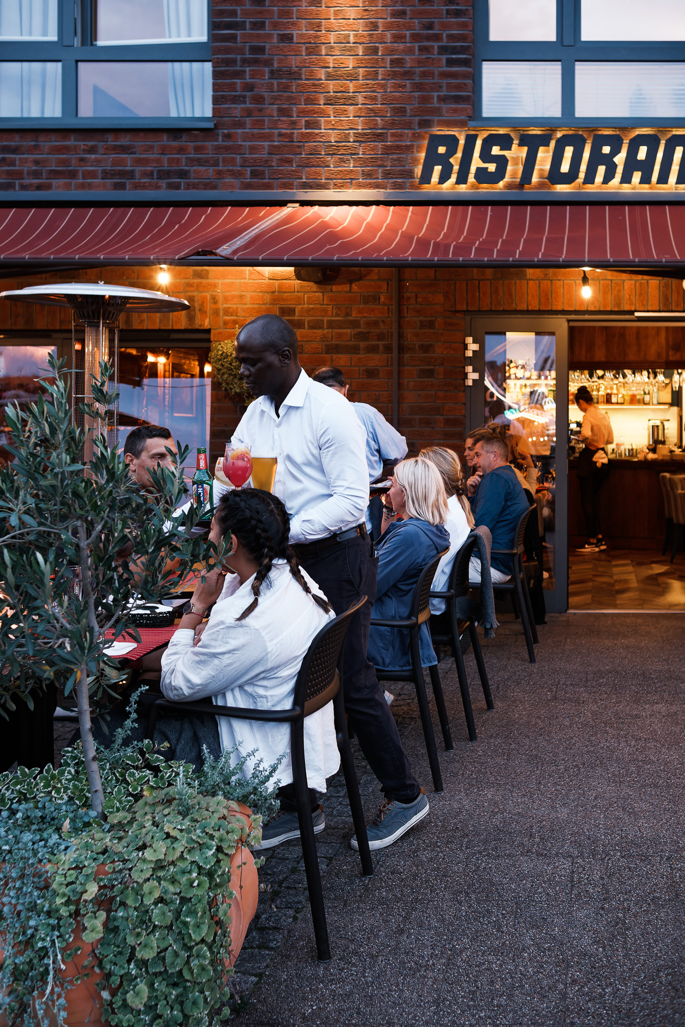 Czarnoskóry kelner podaje napoje gościom siedzącym w ogródku restauracji w Gdańsku o zmierzchu