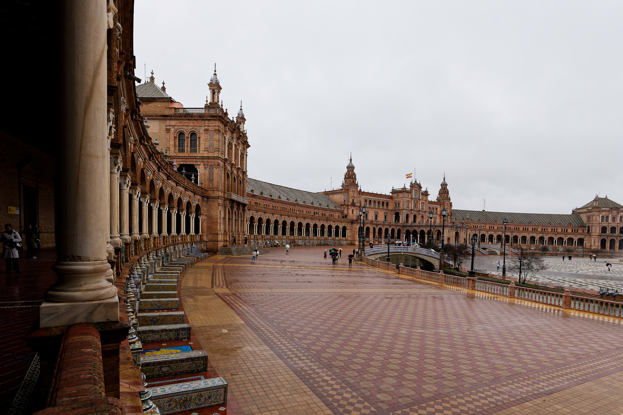 Plac Hiszpański w Sewilli podczas intensywnego deszczu, mokre azulejos i arkady odbijające światło pod ciężkim niebem